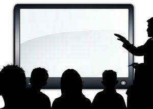 Silloutte of man standing at front of classroom pointing to the large screen. Several people are in the room looking at it.