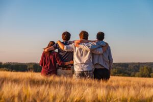 Four teens standing in a field of high grass with their arms around each other looking out at the field and blue sky