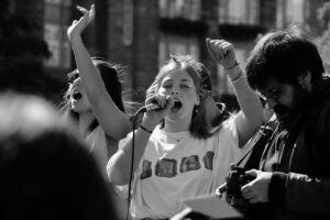 Picture of girl with micraphone at a rally or strike - young people in background raising arms and a man taking pictures