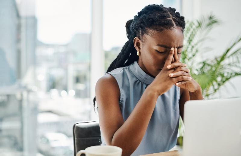 A middle aged, professional woman with long hair sitting at a desk resting her head against her hands and looking downwards. There are large windows behind her and a plant in the background. She has a laptop and coffee on her desk.