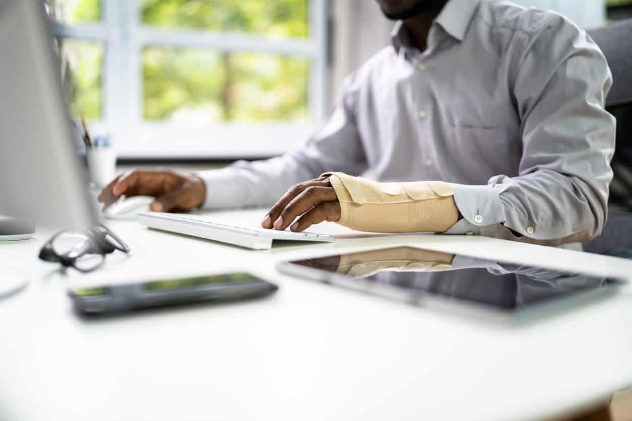 Man working at desk by a window with green trees outside. He has cast on one arm