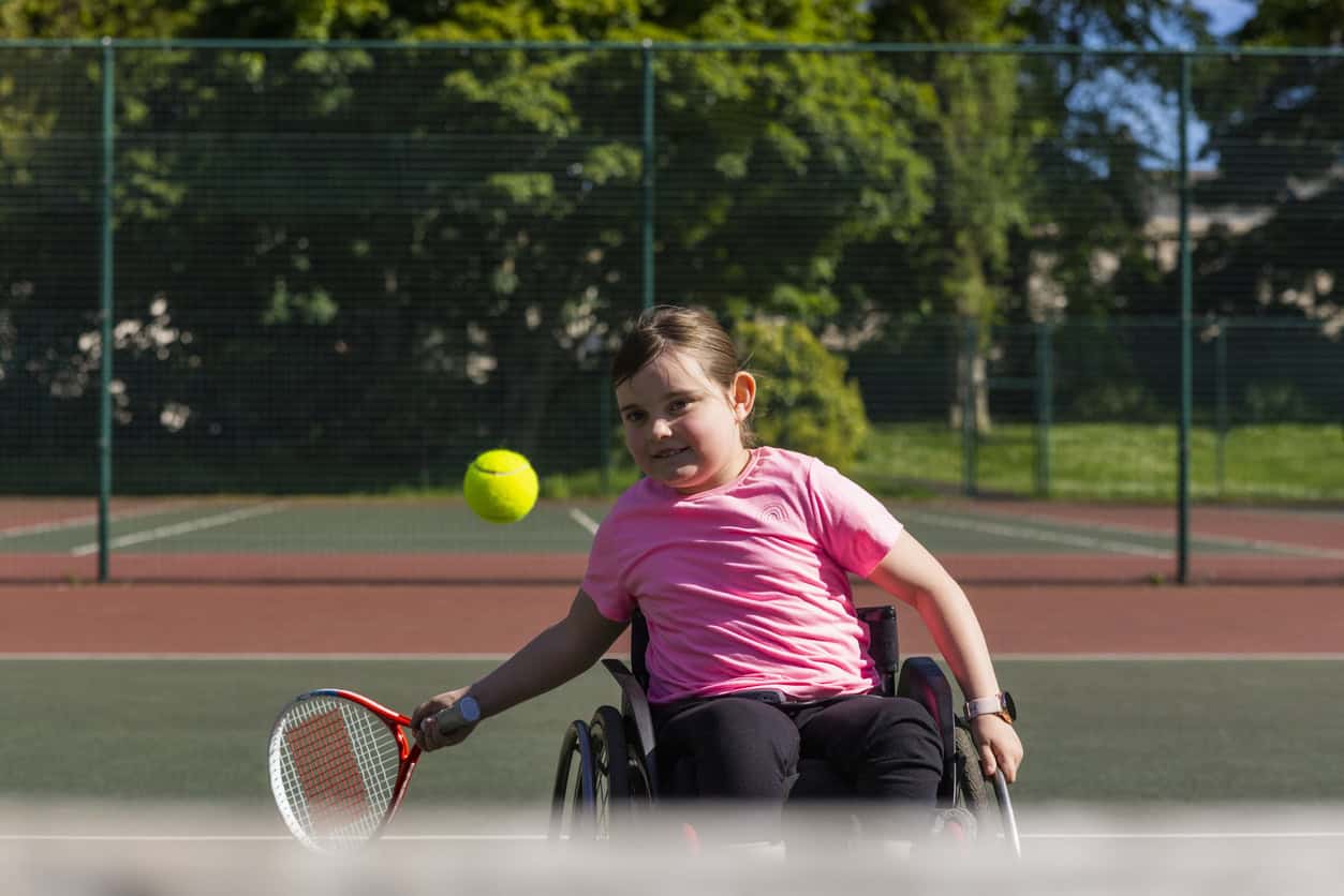 A young girl who is a wheelchair user, playing tennis on a tennis court in a public park in Newcastle upon Tyne, England. She has her racket out, ready to hit the tennis ball on the way to her.
