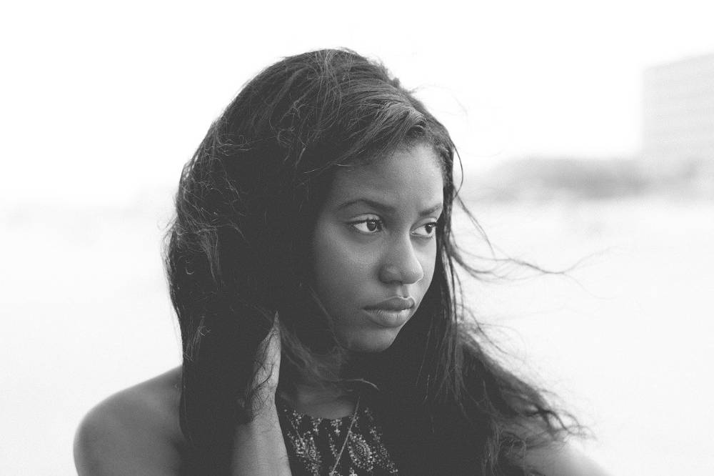 A close up of a young lady, the breeze is blowing her hair. Their is water in the background. She looks sad.