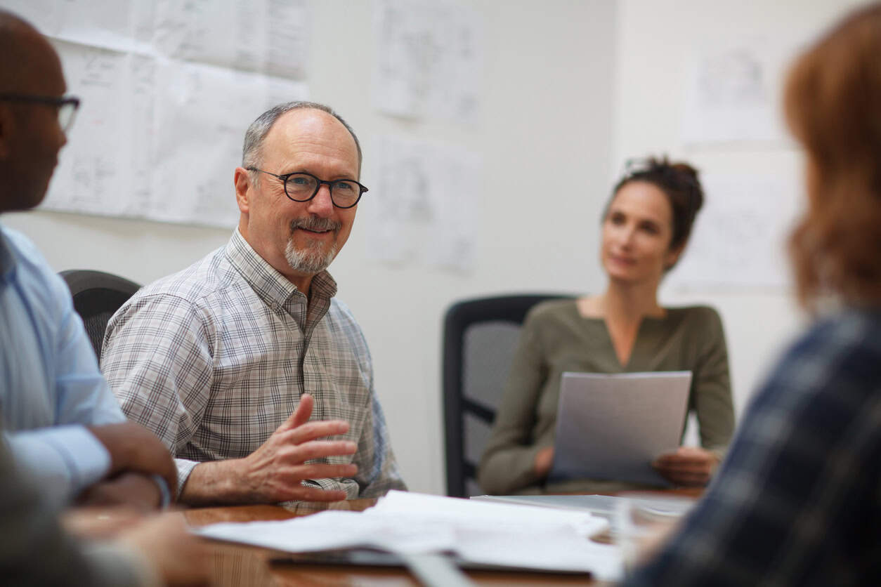 Senior man listens to coworker in meeting with work colleagues while sitting at table in design office