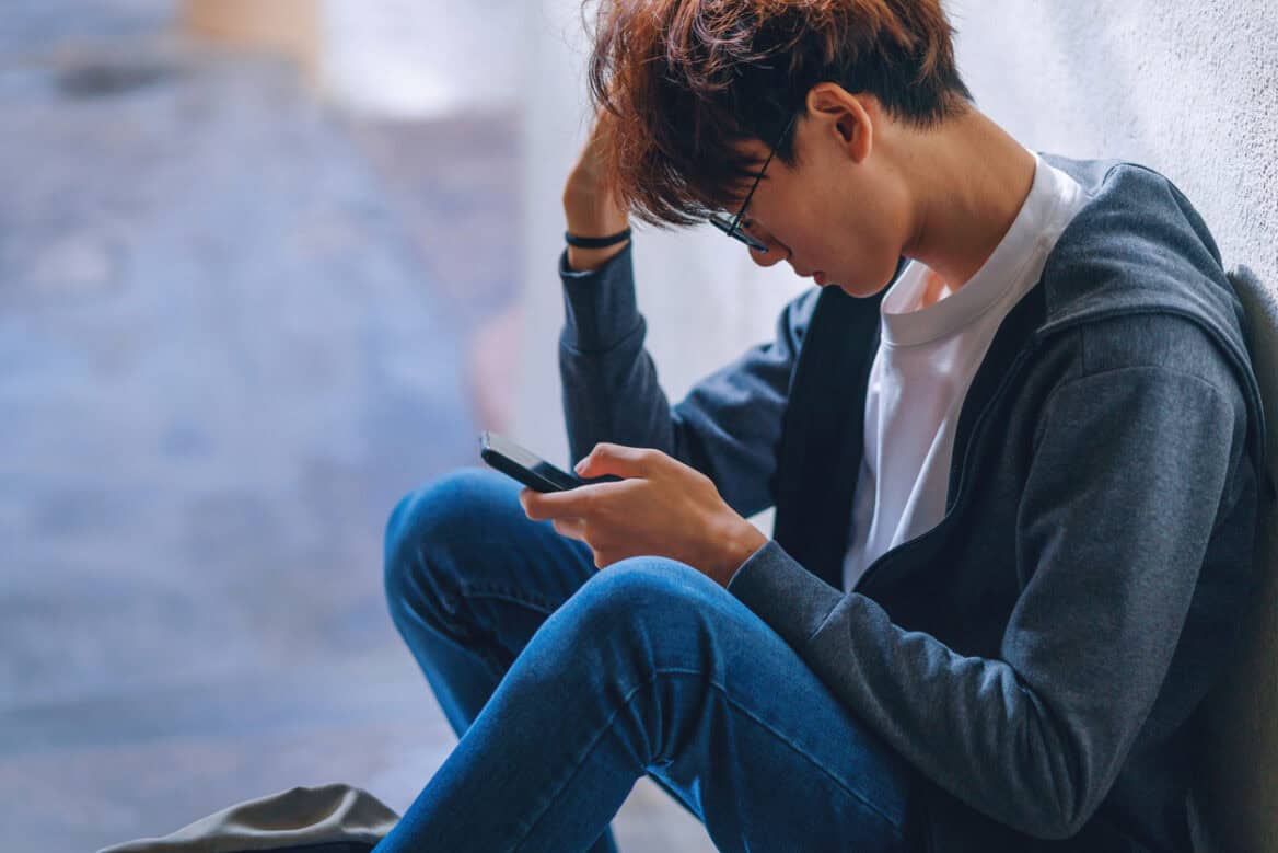 An upset Asian teenage boy student, dressed in casual outfit, sits on a stairway reading on his smartphone with a backpack beside him, while dealing with the issue of cyberbullying.
